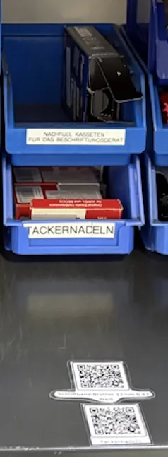 Blaue Sichtlagerkästen mit Tackernadeln und Nachfüllkassetten, davor zwei Nachbestell-QR-Codes auf der Regalkante
