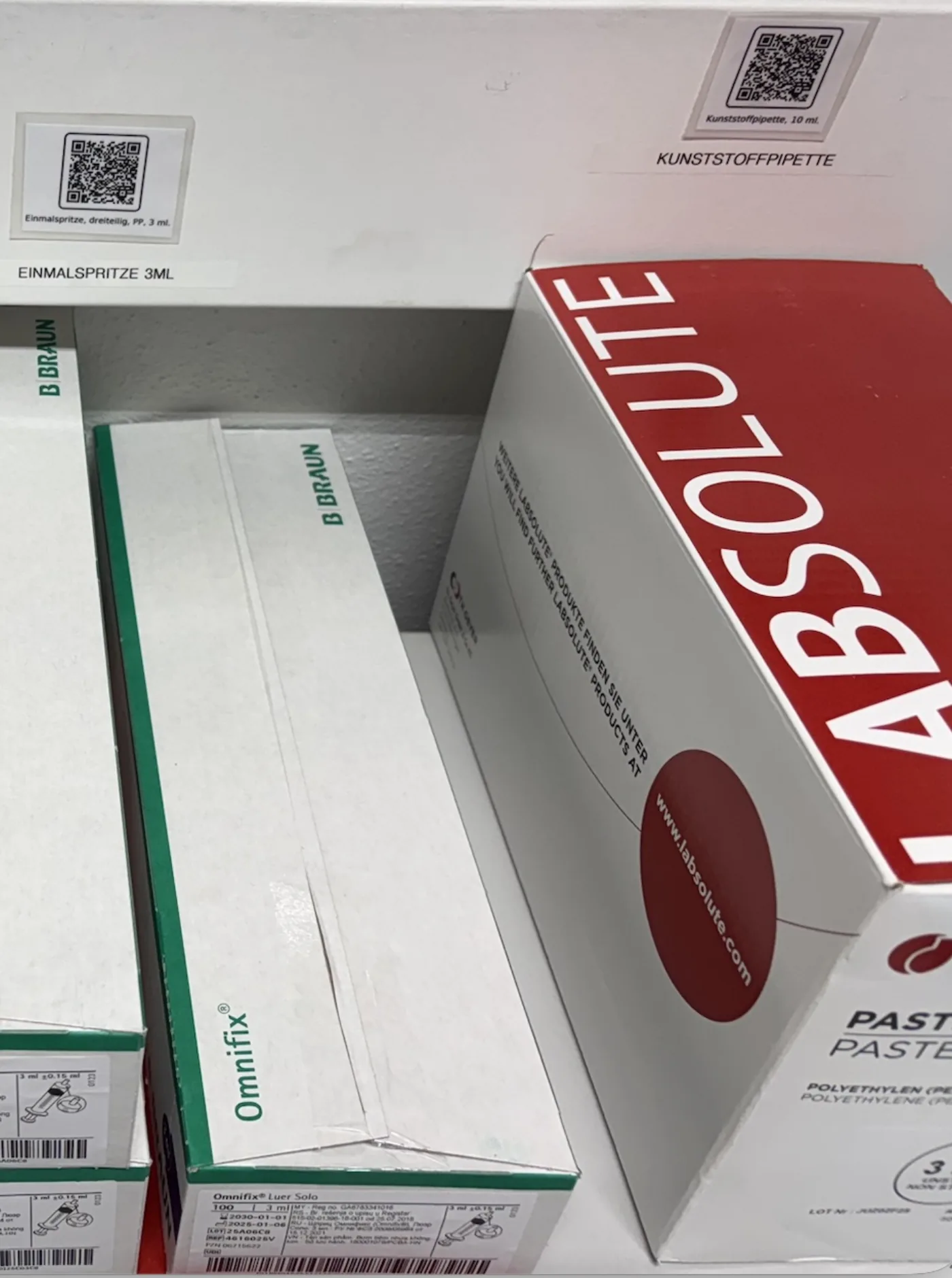 Regalfach mit Einmalspritzen, Kunststoffpipetten und Laborverbrauchsmaterial, QR-Codes mit Artikelbezeichnung am Fachboden