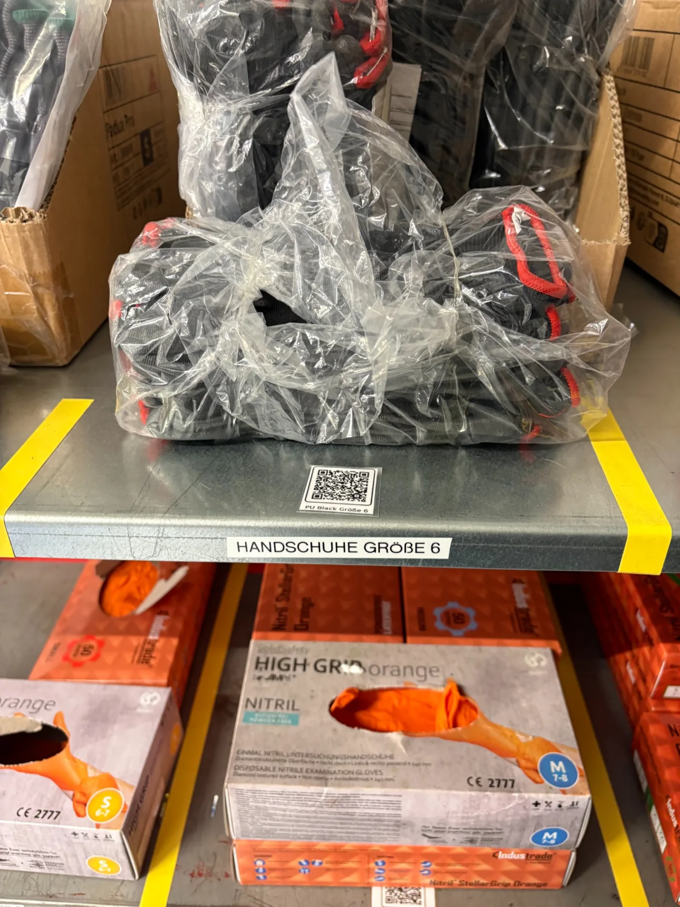 Regalboden mit Arbeitshandschuhen Größe 6 und QR-Code-Etikett, darunter Kartons mit Nitrilhandschuhen