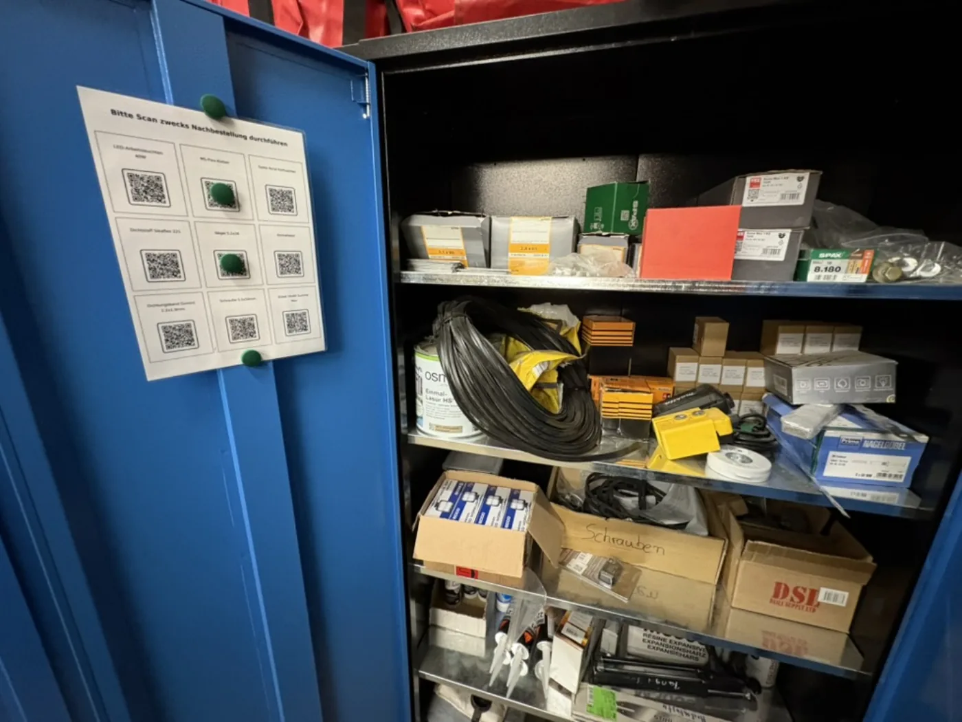 Blauer Materialschrank mit Kleinteilen und Schrauben, an der Tür ein Blatt mit neun Nachbestell-QR-Codes